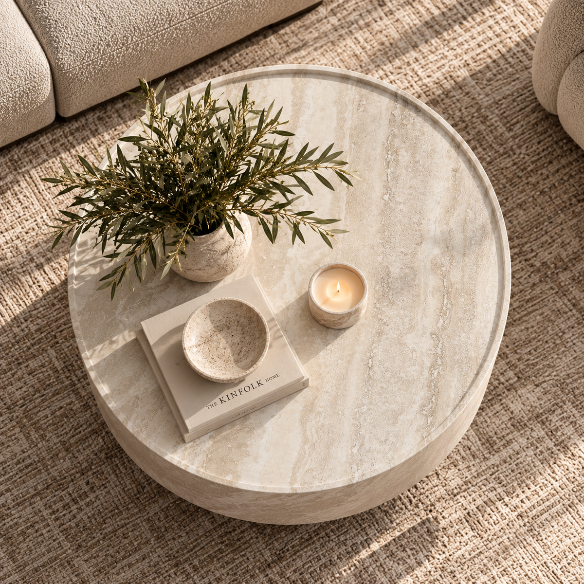 Round light beige stone coffee table in a styled living room setting, decorated with a potted olive branch, a lit candle in a ceramic holder, and a small ceramic bowl on stacked books.