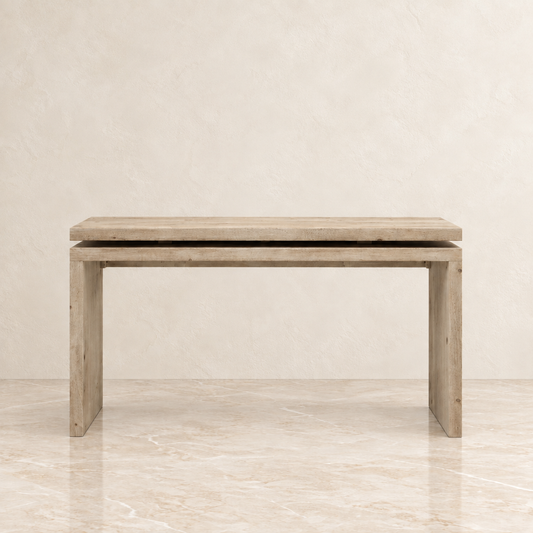 A rustic light beige wooden  console table with a thick rectangular top and four sturdy legs, set against a neutral beige textured wall and marble floor.