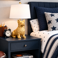 Load image into Gallery viewer, Gold Corgi Lamp with white drum shade on a navy blue nightstand beside a bed with star-patterned bedding.
