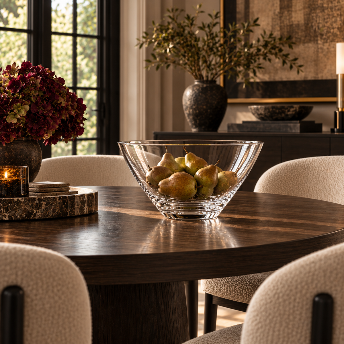 A crystal bowl filled with pears sits on a dark wood dining table in a styled interior with upholstered chairs, a vase of flowers, and warm ambient lighting.