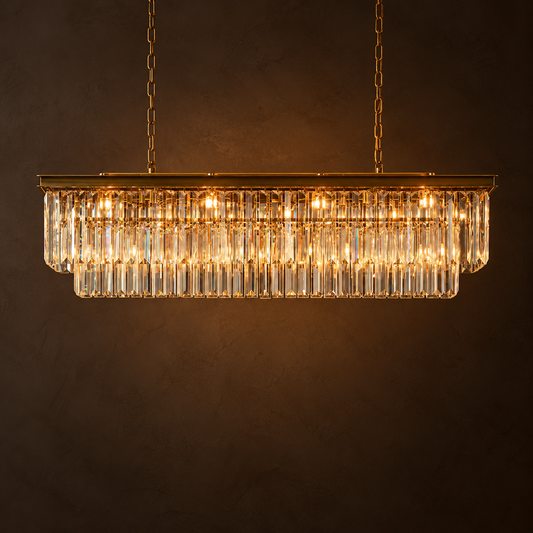 A rectangular chandelier pendant light with a gold-toned metal frame and clear hanging crystal prisms, illuminated against a dark brown background.
