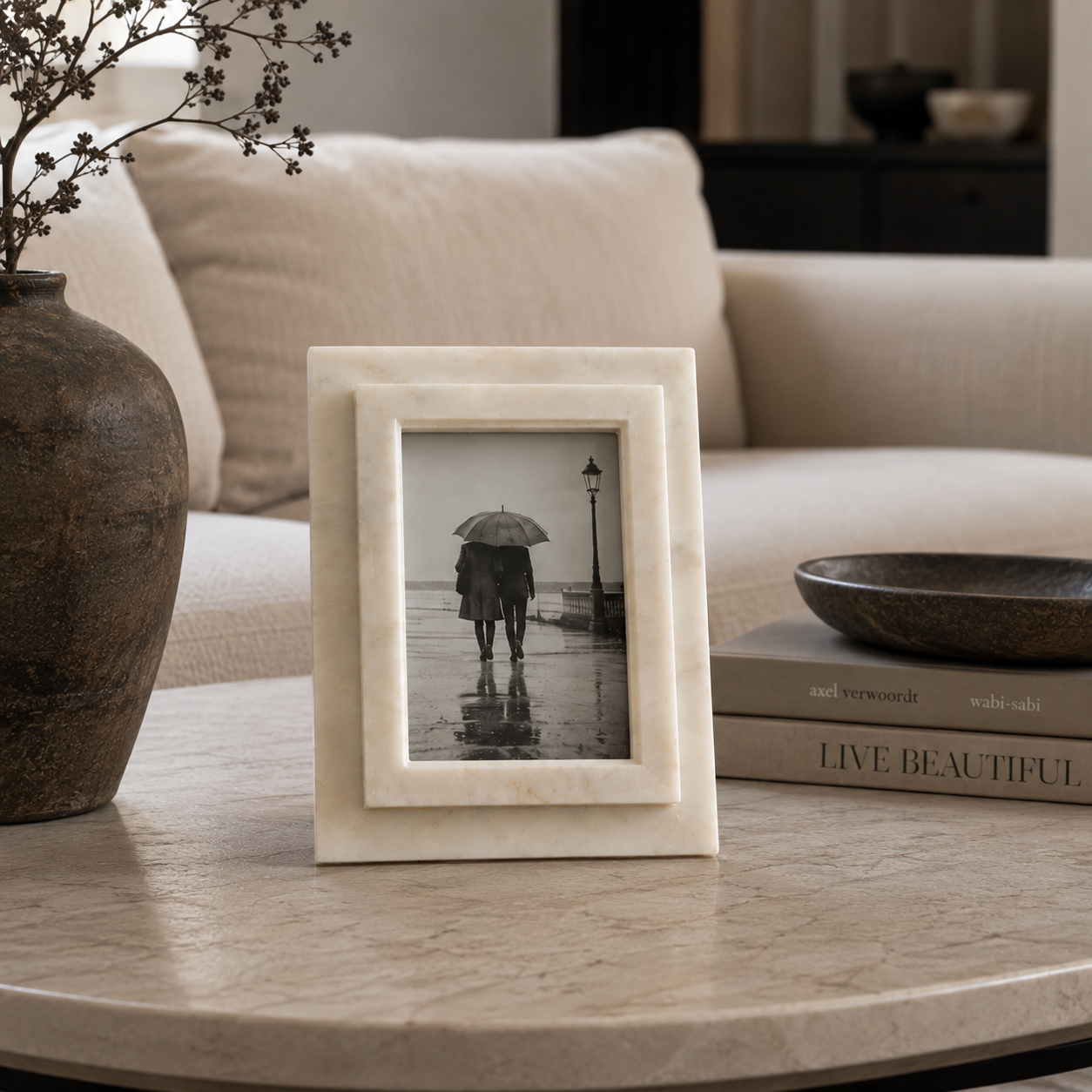 A square cream-colored marble decorative frame with a double-layered border stands on a round stone table in a living room setting. The frame contains a black-and-white photograph of a person holding an umbrella in the rain.