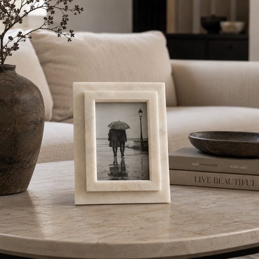 A square cream-colored marble decorative frame with a double-layered border stands on a round stone table in a living room setting. The frame contains a black-and-white photograph of a person holding an umbrella in the rain.