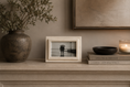 Load image into Gallery viewer, decorative frame holding a black-and-white photograph, styled on a mantel beside a large textured vase with dried branches, books, a bowl, and a lit candle against a warm neutral wall.
