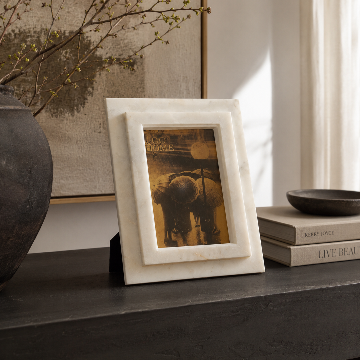 decorative frame with a wide inner mat holding a vintage-style photograph, displayed on a dark tabletop in a styled interior setting with books, a bowl, and dried branches.
