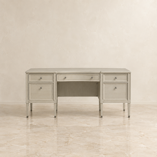 A light gray upholstered writing desk with three drawers, a center drawer, and an open center shelf, shown against a neutral beige textured wall and marble floor.