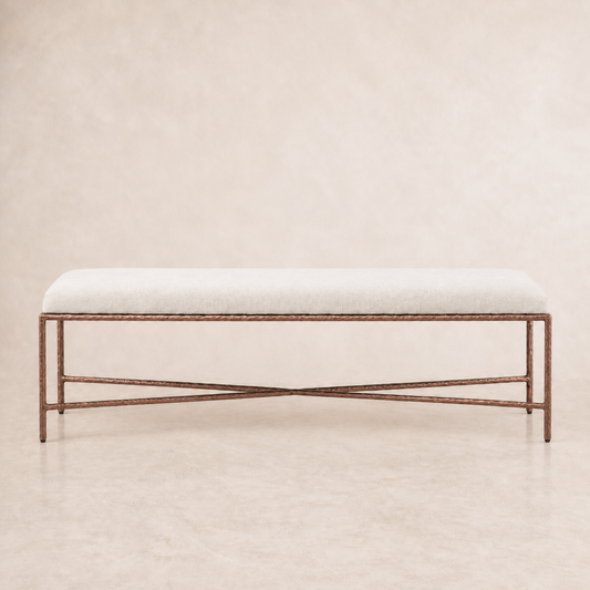 A rectangular upholstered bench with a light beige fabric seat and a dark bronze metal frame featuring crossed lower legs, set against a neutral beige textured background.