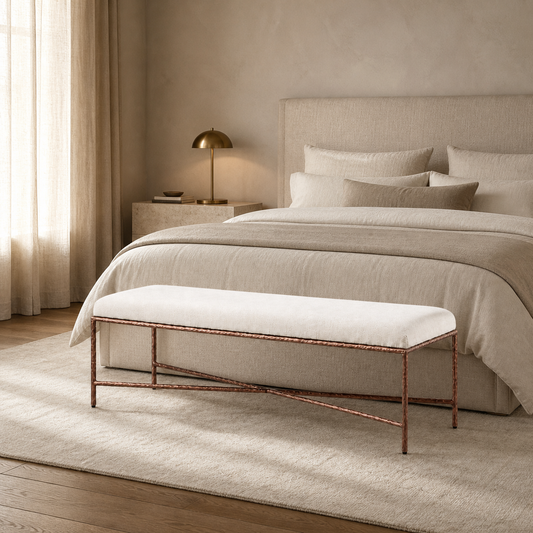 A modern fabric bench with a white cushion and a copper-toned metal frame sits at the foot of a bed in a softly lit neutral bedroom.