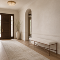 Load image into Gallery viewer, A styled entryway featuring a long cream rug, a white fabric bench with a copper-toned metal frame, a large white ceramic vase with dried branches, and a dark wood double door under a chandelier.
