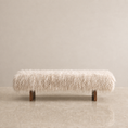 Load image into Gallery viewer, A long rectangular bench upholstered in fluffy cream-colored faux fur or sheepskin, supported by two dark wood legs, set against a minimalist beige wall and floor.
