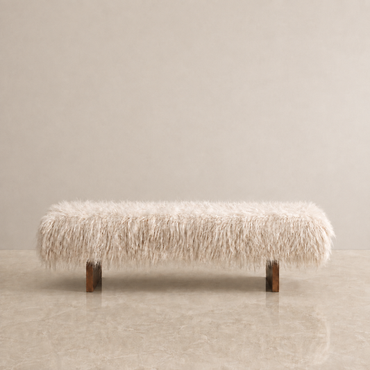 A long rectangular bench upholstered in fluffy cream-colored faux fur or sheepskin, supported by two dark wood legs, set against a minimalist beige wall and floor.