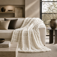 Load image into Gallery viewer, faux fur throw blanket in off-white is draped over a light beige sofa in a modern, sunlit living room. The scene includes neutral-toned pillows, a wooden coffee table with books, and a large window in the background.
