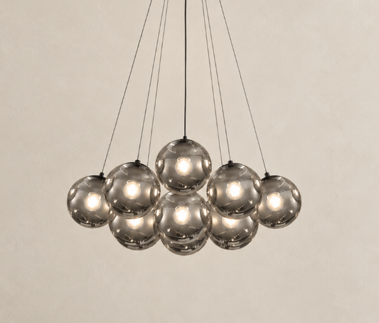 A modern multi-light pendant chandelier with nine reflective smoked glass globes suspended by thin black cords against a warm beige textured wall.