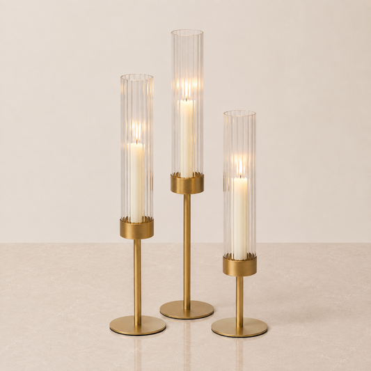 Three gold-toned candlestick holders with clear ribbed glass cylinders and white candles, arranged on a light beige surface against a neutral cream background.