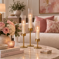 Load image into Gallery viewer, Three gold candlestick holders with clear glass cylinders and lit white taper candles are arranged on a marble coffee table in a styled living room setting.
