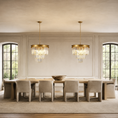 Load image into Gallery viewer, Two matching gold chandelier lights with clear glass crystal drops hang symmetrically above a long dining table and upholstered chairs in a bright, elegant dining room.
