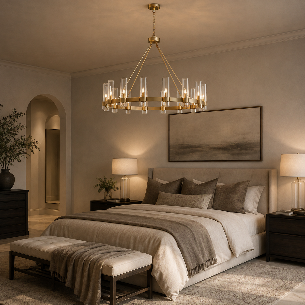 A modern brass chandelier with clear glass cylindrical tubes hangs over a bed in a styled bedroom. The room features neutral beige walls, a beige upholstered bed with gray and beige bedding, matching pillows, a bench at the foot of the bed, and dark wood nightstands with lit lamps.