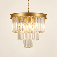 Load image into Gallery viewer, A modern tiered chandelier with a brushed gold metal frame and clear glass cylindrical prisms, illuminated against a warm off-white background.
