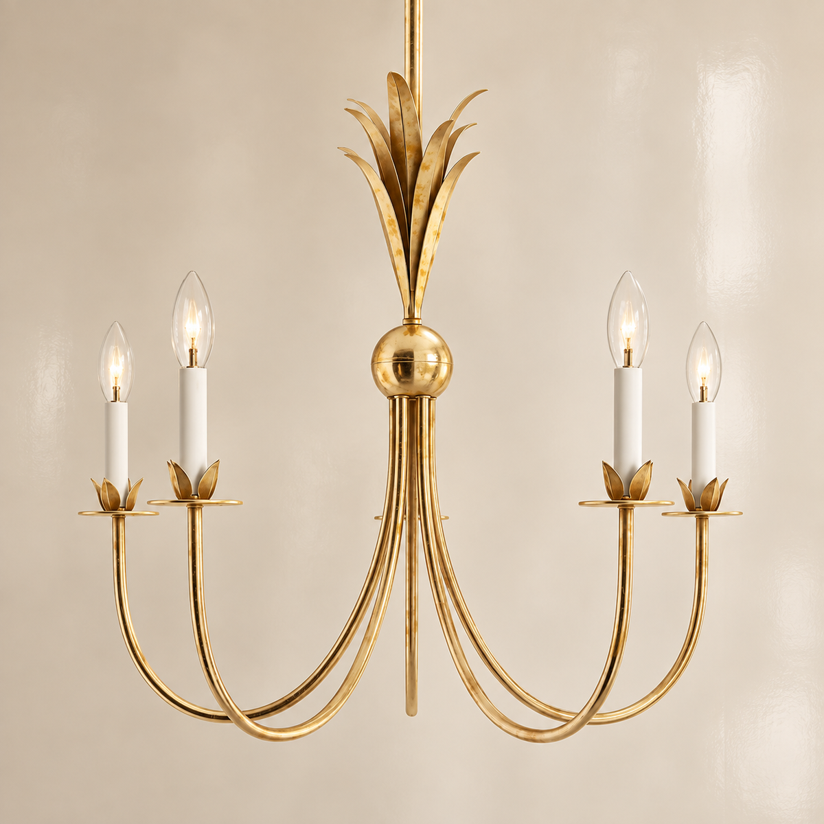 A five-light gold chandelier with curved candle-style arms, a central round finial, and a leafy top ornament, shown against a plain beige wall.