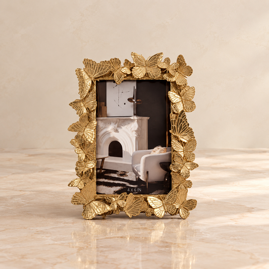 A rectangular gold picture frame with an ornate border made of metallic gold leaves, standing on a light beige marble surface against a matching beige wall. The frame contains a sample photograph of a fireplace.