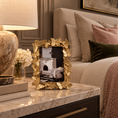 Load image into Gallery viewer, A decorative gold picture frame with a leaf border sits on a marble nightstand beside a lit lamp and flowers, with a bed in the background.
