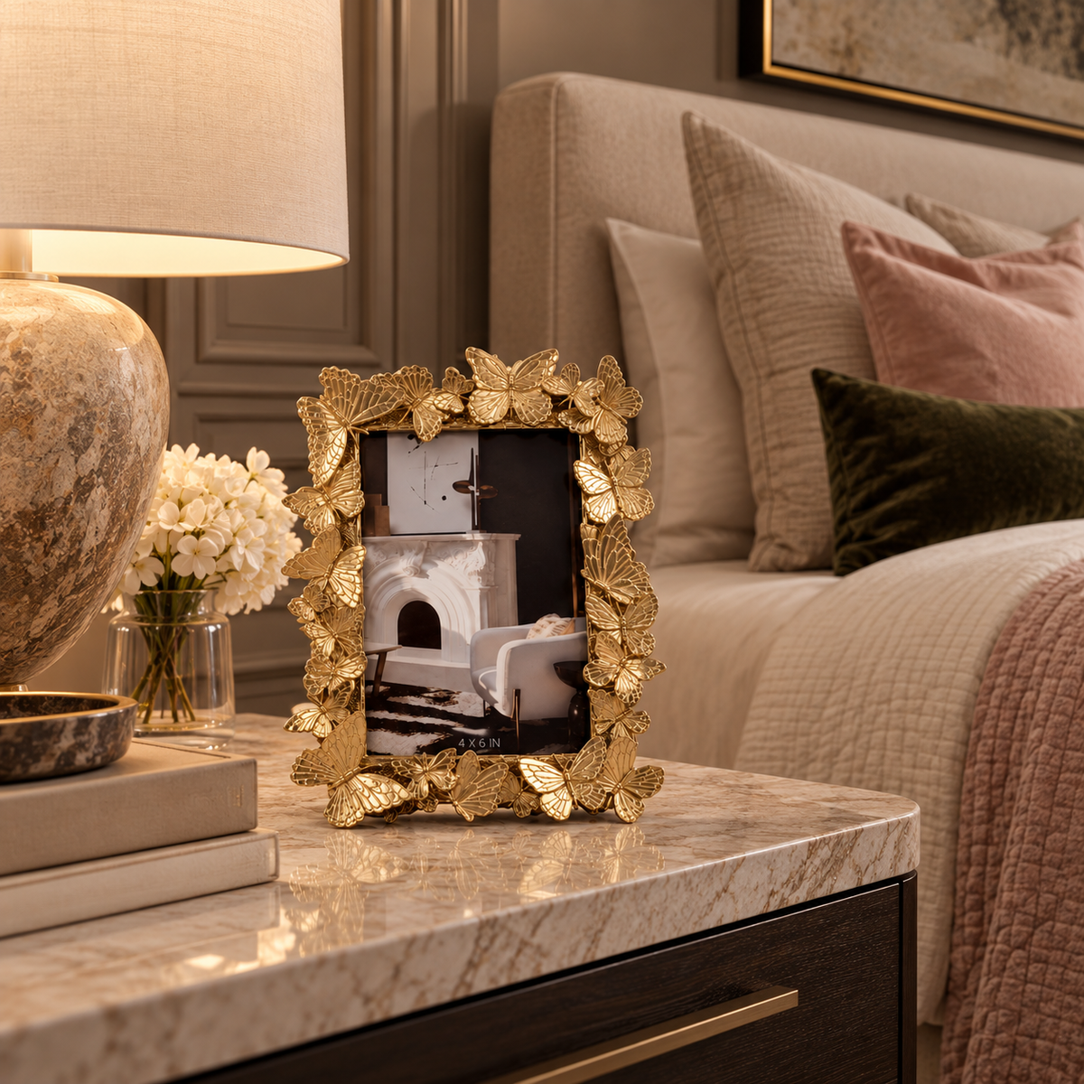 A decorative gold picture frame with a leaf border sits on a marble nightstand beside a lit lamp and flowers, with a bed in the background.