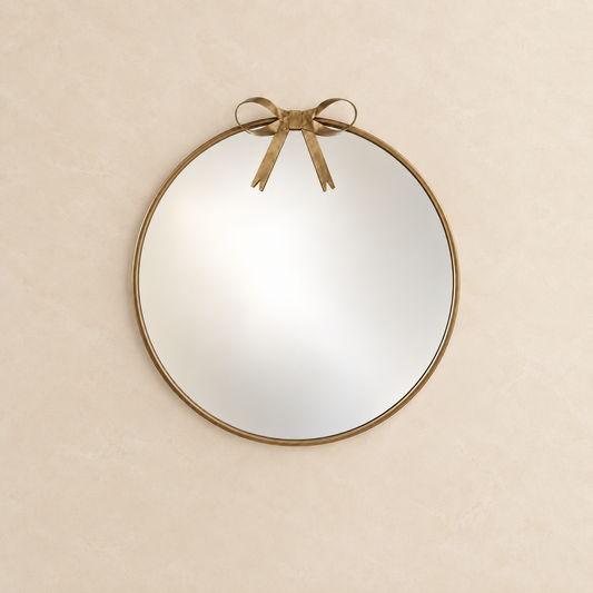 gold roundwall mirror with a distressed brass frame and a matching bow detail at the top, mounted on a textured beige wall