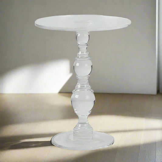 A small, round ACRYLIC END TABLE with an intricately designed central support from House of Effy stands on a light wooden floor in a brightly lit room, adding a modern touch to the space.