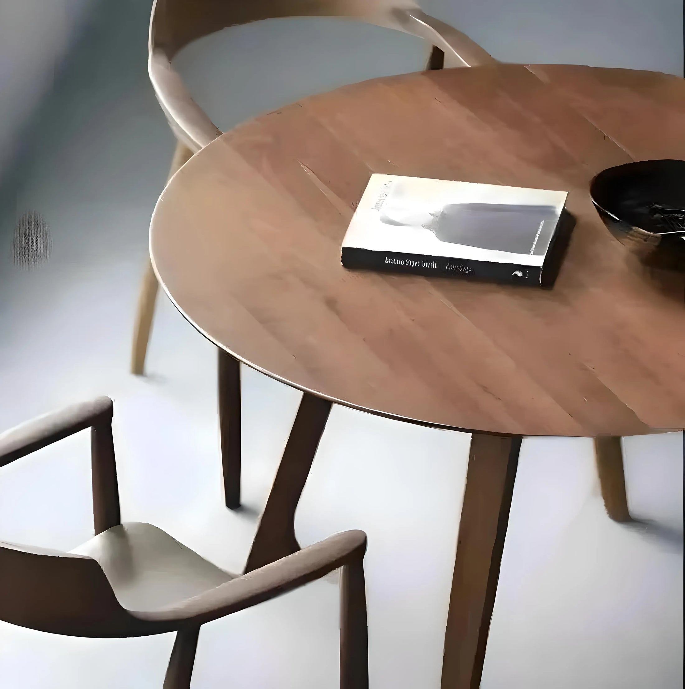 A modern minimalist setup with a round House of Effy pine wood dining table, two chairs, a book titled "excess isn’t enough", and a round dark tray.