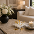Load image into Gallery viewer, A styled living room scene showing a marble-top table with a white marble tray holding several gold-toned ring stands, alongside a black vase of white flowers, a lit candle, and a cream sofa in the background.
