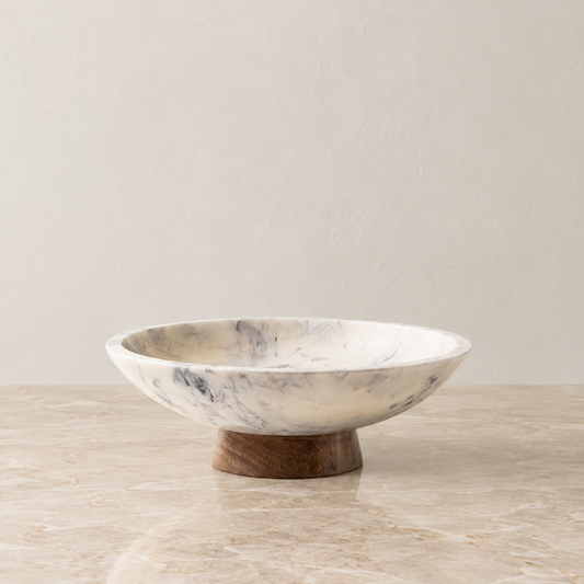 A round marble pedestal bowl with a white and gray marbled pattern, resting on a small wooden base over a light beige marble surface.