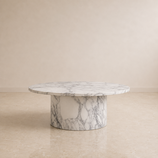 A round marble pedestal coffee table with a white and gray veined pattern, set against a minimalist beige wall and tiled floor.