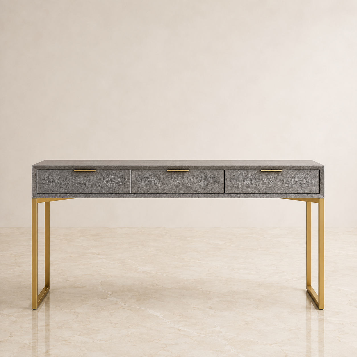 A modern console table with a gray shagreen top and three drawers, supported by a slim gold metal frame, set against a neutral beige background.