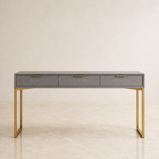A modern console table with a gray shagreen top and three drawers, supported by a slim gold metal frame, set against a neutral beige background.
