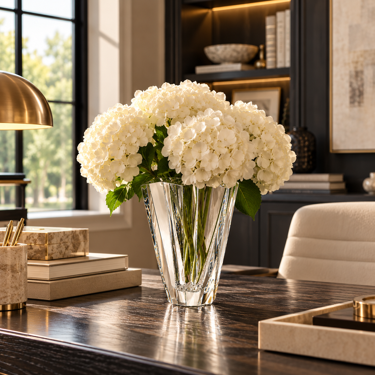 A crystal vase of white hydrangeas, displayed on a dark wood desk in a modern, upscale home office.