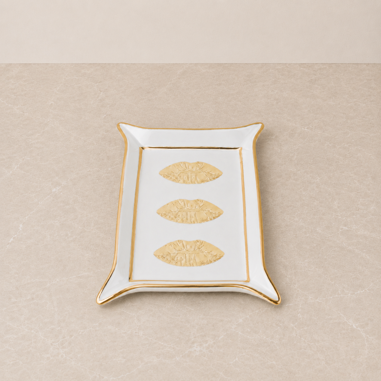 A rectangular white ceramic tray with a gold rim, featuring three embossed gold lips motifs down the center, set against a neutral beige textured background.