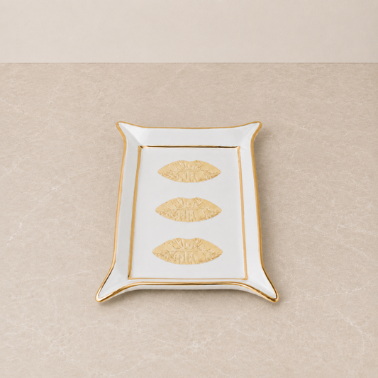 A rectangular white ceramic tray with a gold rim, featuring three embossed gold lips motifs down the center, set against a neutral beige textured background.