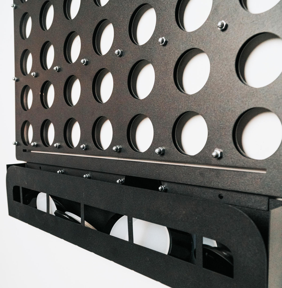 Close-up of black metal life size wall-mounted Four in a Row game with round slots and disc storage tray on white background