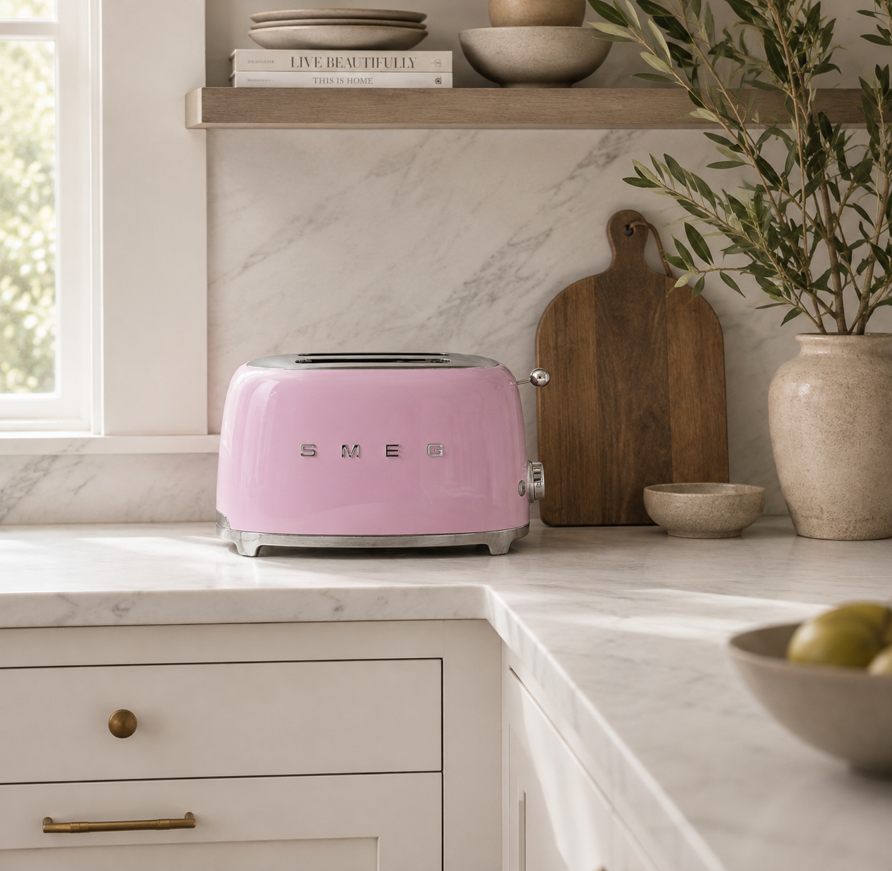 pink retro toaster sits on a white marble kitchen countertop, surrounded by wooden cutting boards, ceramic bowls, and a potted olive branch in a bright, modern kitchen.