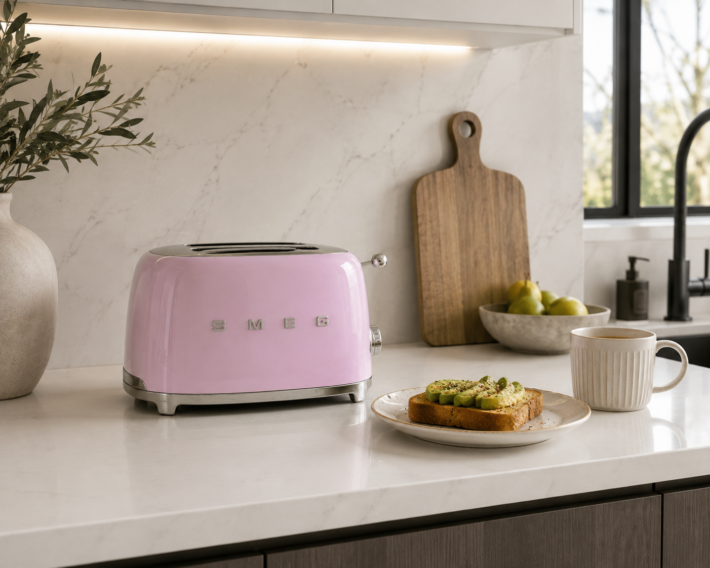 pink retro toaster sits on a white kitchen countertop next to a plate of avocado toast, a white mug, a wooden cutting board, and a vase with greenery in a bright, modern kitchen.