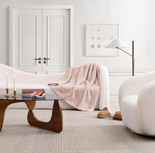 Pink Throw Blanket by UGG draped over a cured sofa in a living room next to a floor lamp and across from a coffee table and a boucle swivel chair