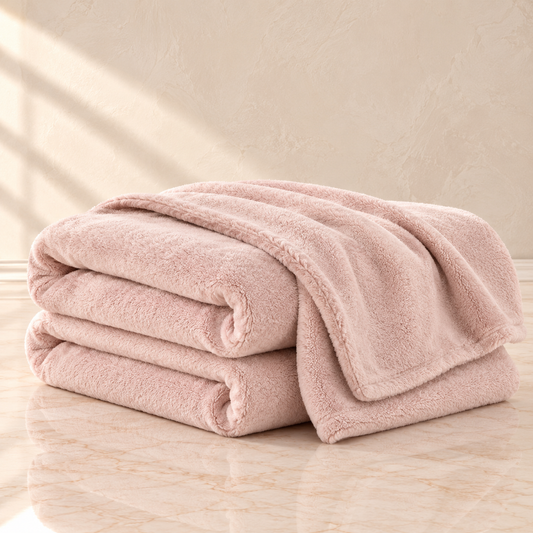A stack of three folded plush throw blankets in a soft dusty pink color, set on a light beige marble surface against a textured beige wall with soft window shadow light.