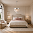Load image into Gallery viewer, A luxury bedroom features a large bed dressed in cream and blush pink linens, flanked by matching nightstands and lamps, with a round crystal chandelier hanging above in a warm, beige-paneled room.
