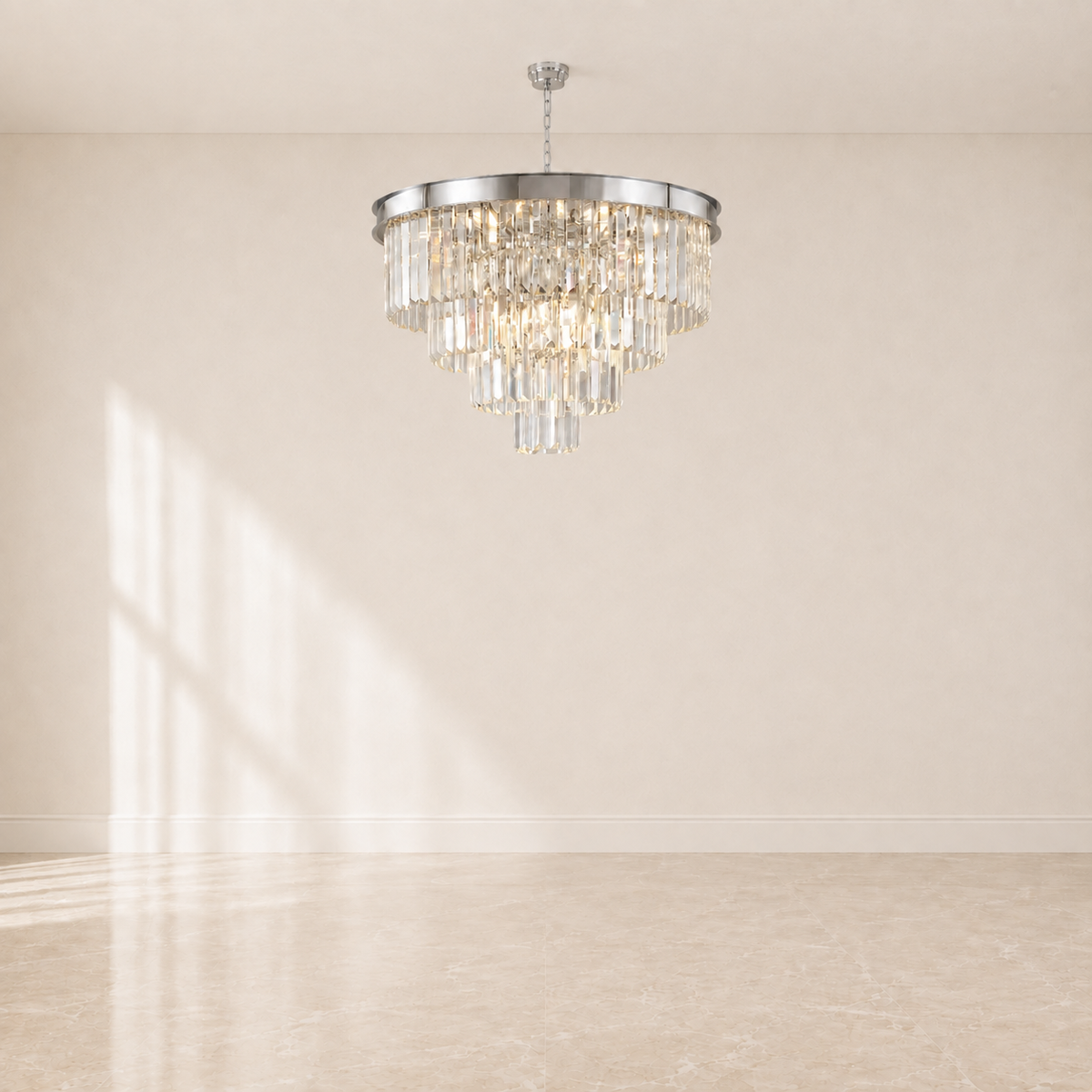 A tiered crystal chandelier with a polished silver-toned metal frame and hanging clear glass prisms, suspended from the ceiling in a minimalist room with beige walls and floor.