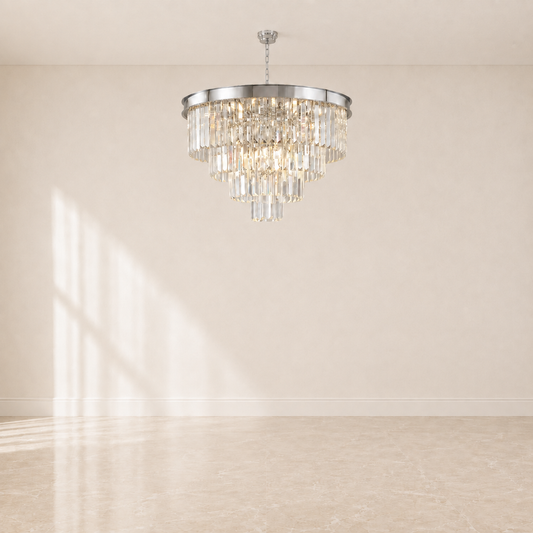 A tiered crystal chandelier with a polished silver-toned metal frame and hanging clear glass prisms, suspended from the ceiling in a minimalist room with beige walls and floor.