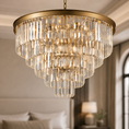 Load image into Gallery viewer, A large tiered round gold crystal chandelier with a brushed gold metal frame and hanging clear glass prisms, shown illuminated in a softly blurred bedroom setting.
