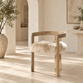 Load image into Gallery viewer, A wooden skeepskin chair with a curved backrest and a fluffy cream sheepskin seat cushion, set in a warm, sunlit minimalist interior with beige stone floors and arches.
