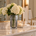 Load image into Gallery viewer, White hydrangea flowers arranged in a tall ribbed clear glass vase on a marble bathroom countertop beside a gold faucet, soap dispenser, and lit candle.
