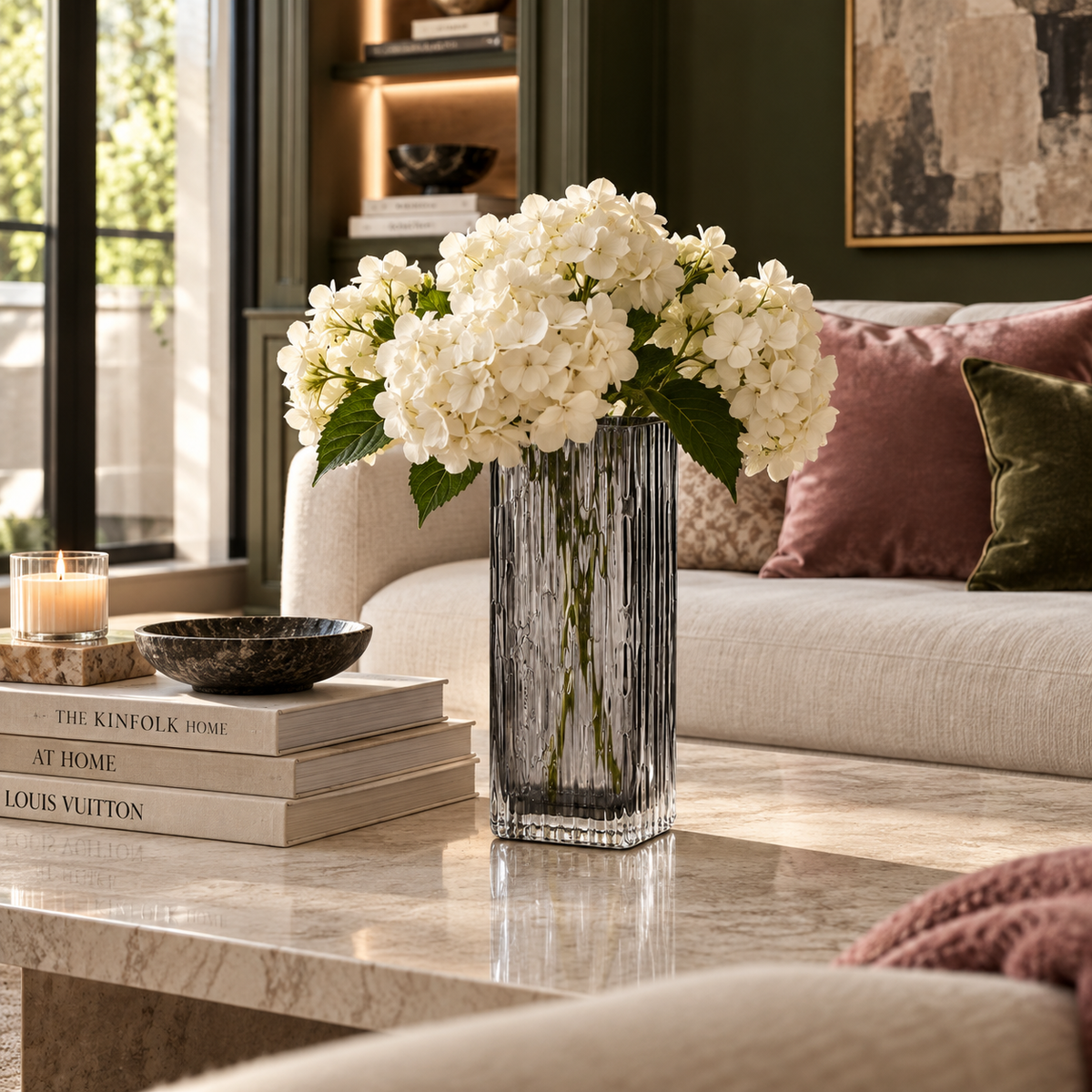 A tall ribbed clear glass vase filled with white hydrangea blooms, placed on a marble coffee table in a styled living room with a sofa, pillows, books, and candles.