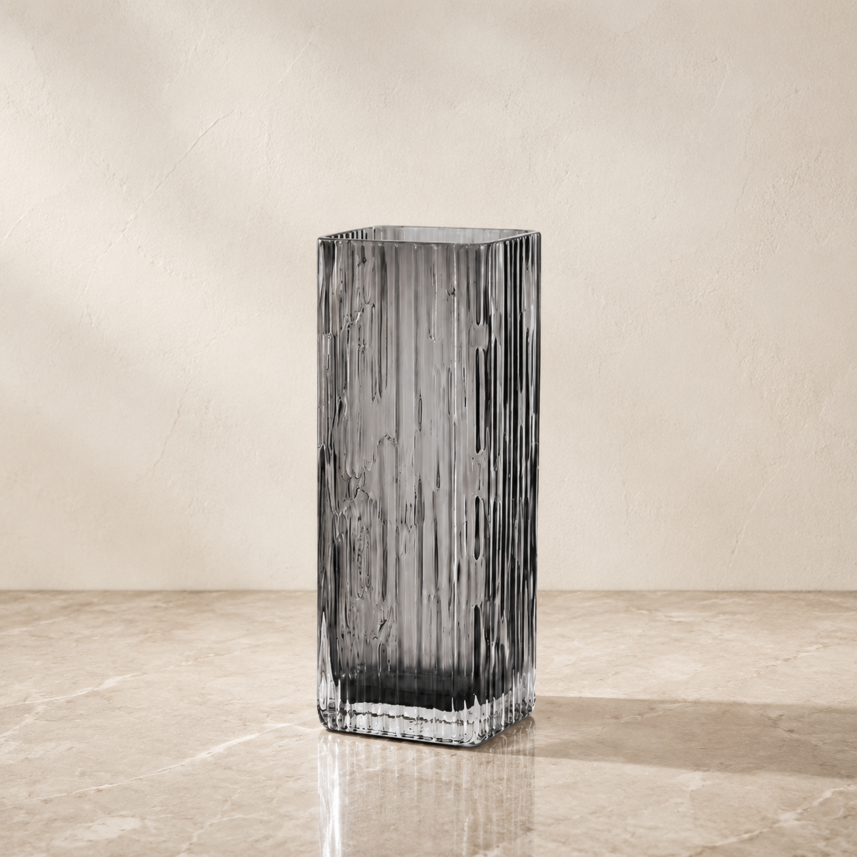 A tall, rectangular ribbed glass vase in a smoky translucent gray color, standing on a beige marble surface against a textured beige wall.